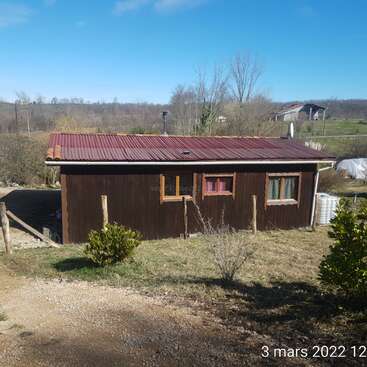 The image depicts a small, one-story house with a brown exterior and red roof, situated in a rural setting on a sunny day.