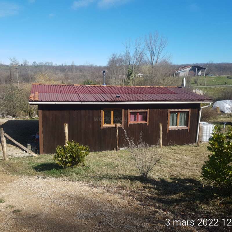 The image depicts a small, one-story house with a brown exterior and red roof, situated in a rural setting on a sunny day.