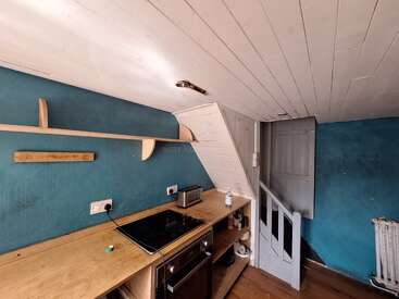 This image shows a small, rustic kitchen with blue walls, white slanted ceiling, wooden shelves, a stove, toaster, oven, and open shelving. There’s a stairway.