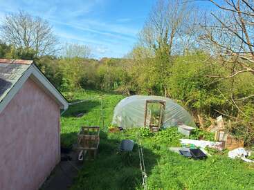 A rural backyard with a pink building, green grass, a plastic greenhouse, wooden shelves, gardening tools, and scattered supplies surrounded by trees under a blue sky.