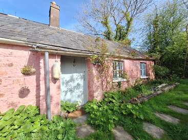 A charming pink cottage with a slate roof, white door, and blue-framed windows, surrounded by lush plants, greenery, sunlight, and a tranquil garden.