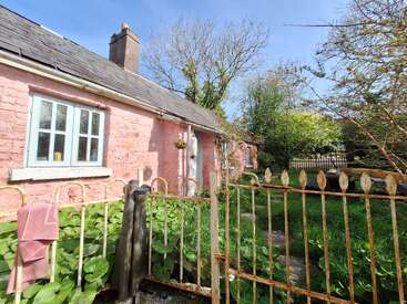 A charming pink cottage with a slate roof sits behind a rusty iron gate, surrounded by lush greenery, overgrown plants, and dappled sunlight on a clear day.