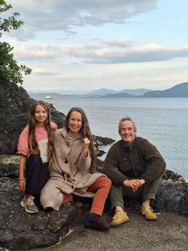 A smiling family of three sits on rocky shore beside calm water, with distant mountains, a cruise ship, and cloudy sky in the peaceful background.