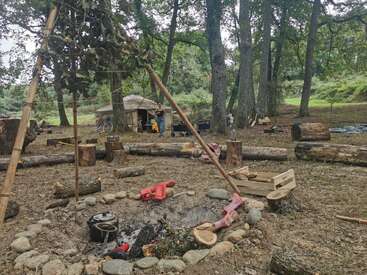 A rustic outdoor campsite in a forest features a central fire pit with cooking equipment, wooden logs for seating, a tent, and people setting up in the background.