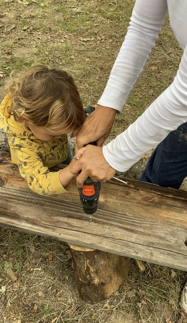 A child and an adult work together outdoors, using a power drill on a wooden plank. The scene conveys learning, teamwork, and hands-on exploration in nature.