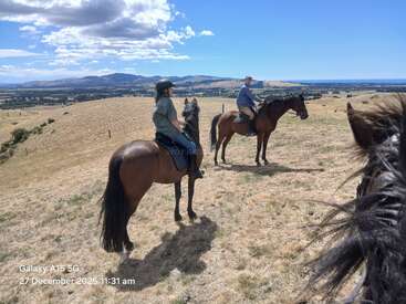 Deux personnes à cheval traversent des collines sèches et vallonnées sous un ciel bleu. La tête d'un autre cheval apparaît au premier plan. Des montagnes et des plaines lointaines sont visibles.