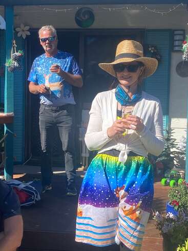 Une femme portant un chapeau et des lunettes de soleil sourit, vêtue d'une jupe colorée sur le thème de l'hiver. Derrière elle, un homme vêtu d'une chemise de renne festive boit à l'extérieur.