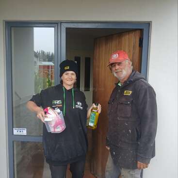 Deux personnes se tiennent devant une porte ouverte, en souriant. L'une tient un récipient en plastique et une bouteille. Tous deux portent des bonnets et des sweats à capuche, et ont l'air décontractés et amicaux.