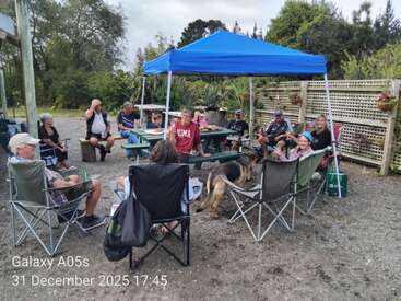 Um grupo de pessoas se junta ao ar livre sob um toldo azul, sentadas em cadeiras e bancos, curtindo uma conversa e comida, com a companhia de um cachorro, na véspera de Ano Novo.