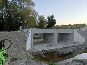 A person stands on a newly constructed concrete culvert over a small creek, surrounded by dirt, trees, and construction equipment on a clear day.