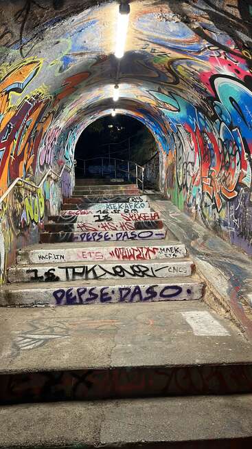 Un escalier à l'intérieur d'un tunnel recouvert de graffitis et de tags colorés, éclairé par des lumières vives au plafond, menant vers le haut, dans l'obscurité ou peut-être vers une sortie.