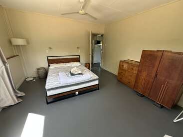 Une chambre simple avec un lit double, une armoire en bois, une commode, un lampadaire, un ventilateur de plafond et des murs clairs. La lumière du soleil pénètre par les fenêtres équipées de rideaux.