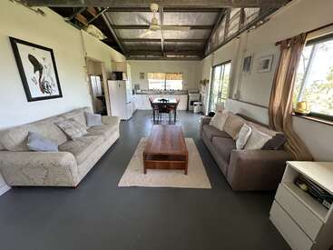 Cet espace de vie ouvert et confortable comprend deux canapés, une table basse en bois, de grandes fenêtres, une petite cuisine, une table à manger, de hauts plafonds et une décoration minimaliste et rustique.
