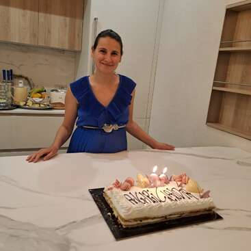 A woman in a blue dress stands behind a white counter with a cake decorated with candles and sweets. The kitchen background features fruit and utensils.