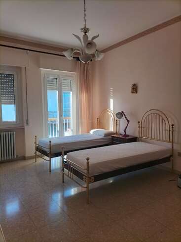 Das ist ein helles Schlafzimmer mit zwei Einzelbetten, goldenen Bilderrahmen, einem Kronleuchter, einer Schreibtischlampe, Marmorboden, Balkontüren und minimaler Wanddekoration.