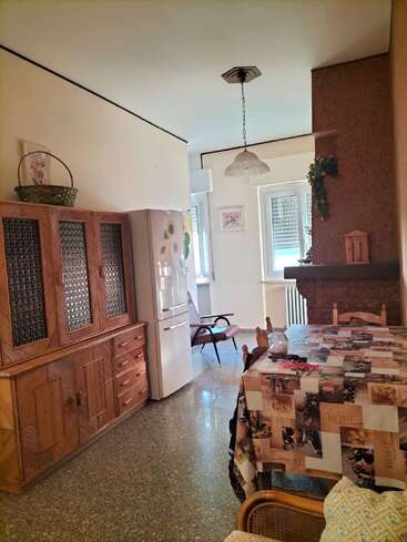 Das Bild zeigt ein gemütliches Esszimmer mit einer gemusterten Tischdecke, Holzmöbeln, einem Kühlschrank, einem Sessel, Wanddekorationen, einem großen Schrank und hellem Tageslicht.