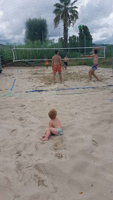 Ein kleines Kind sitzt im Sand und guckt einer Gruppe von vier Leuten zu, die auf einem Sandplatz Beachvolleyball spielen, umgeben von viel Grün und Bäumen.