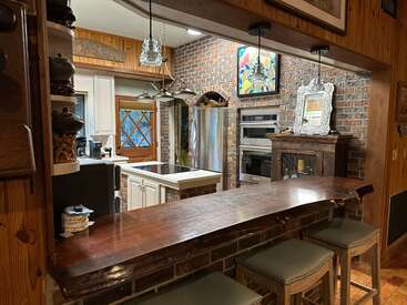 This cozy kitchen features a rustic wooden bar, brick walls, pendant lighting, stainless steel appliances, a decorative mirror, and art, creating a warm, inviting atmosphere.