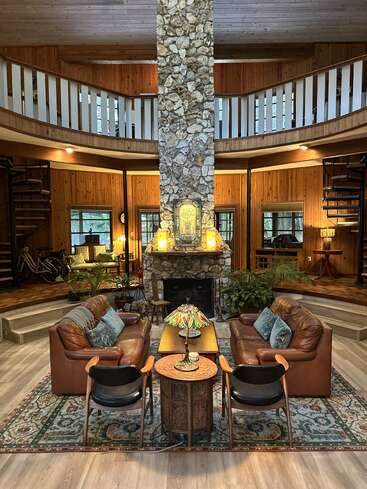 This image shows a cozy, circular living room featuring a tall stone fireplace, leather sofas, wooden walls, spiral staircases, decorative lamps, plants, and a balcony.
