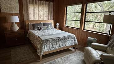 This cozy bedroom features wood-paneled walls, large windows with forest views, a queen bed with gray bedding, nightstands, a white armchair, and soft, neutral decor.