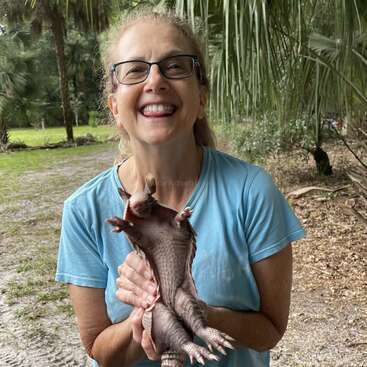 Una mujer sonriente, con gafas y una camiseta azul, está de pie al aire libre, sosteniendo alegremente un pequeño armadillo boca abajo, rodeada de frondosos árboles y plantas.