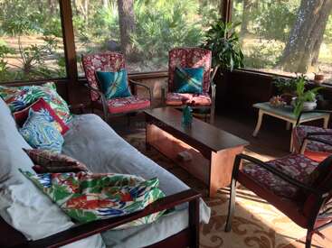 A cozy sunroom features floral patterned chairs, a cushioned sofa with colorful pillows, wooden tables, potted plants, and large windows offering peaceful, lush outdoor garden views.