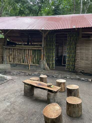 Das Bild zeigt einen rustikalen Sitzbereich im Freien mit einer Holzbank und Baumstümpfen vor einem kleinen, offenen Gebäude mit einem Wellblechdach.