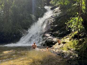 Three people enjoy a waterfall in a lush forest setting. Sunlight filters through the trees, illuminating the cascading water and vibrant green surroundings for a tranquil scene.