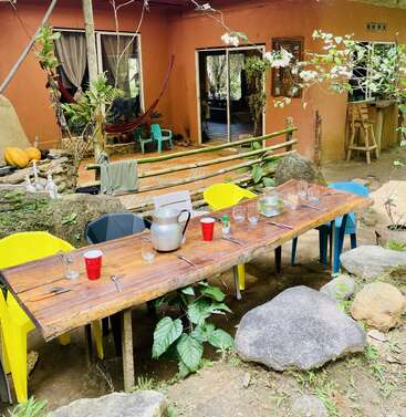 A rustic outdoor dining table with colorful chairs is set for a meal. Hammock, plants, and rocks create a cozy, inviting, natural atmosphere by a house.
