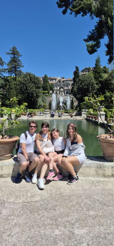 A family sits in front of a beautiful fountain and pond, surrounded by lush greenery, vibrant plants, and classical architecture beneath a clear blue sky.