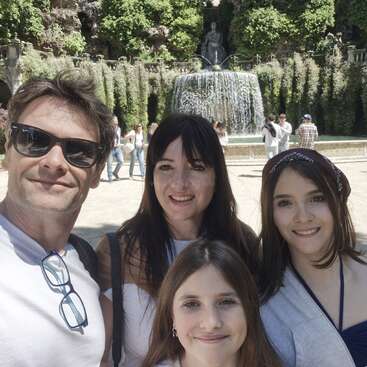 A smiling family of four takes a selfie in front of a beautiful fountain and lush greenery, with several people enjoying the scenic garden in the background.