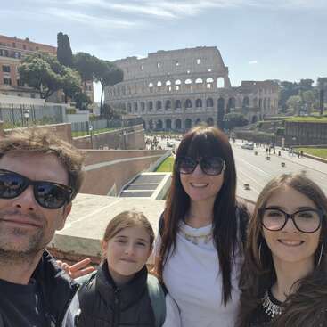 Una familia de cuatro miembros posa para una selfie bajo el sol con el emblemático Coliseo de Roma al fondo, todos sonrientes y disfrutando de su aventura turística.