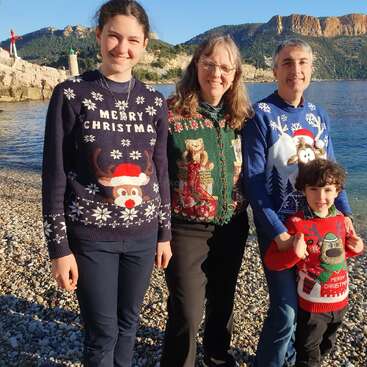 The image shows a family of four standing on a rocky beach, wearing festive Christmas sweaters, with a body of water and a mountainous landscape in the background.