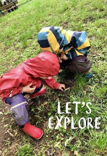 Two children in colorful rain gear crouch on grassy ground, curiously examining something small on the earth. The words “LET’S EXPLORE” are written beside them.
