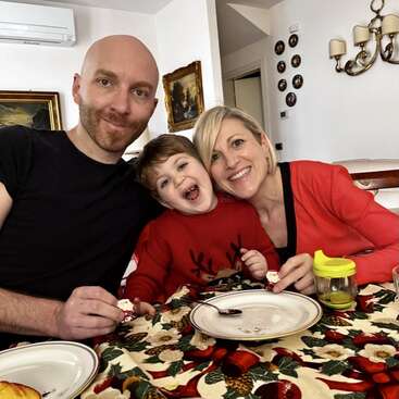 A happy family sits together at a festive table, smiling for the camera. The child wears a red sweater; everyone looks joyful during a holiday meal.