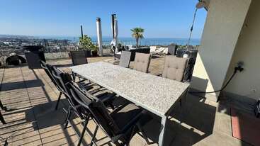 Um espaçoso terraço ao ar livre apresenta uma grande mesa de pedra cercada por cadeiras, espreguiçadeiras, vasos de plantas e oferece uma vista deslumbrante do oceano sob um céu azul claro.