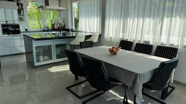 Esta imagem mostra uma cozinha moderna e uma área de jantar com uma mesa grande, seis cadeiras pretas, banquetas brancas, uma ilha e a luz do sol atravessando as cortinas.