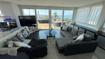 Esta moderna sala de estar possui dois sofás cinza, uma mesa de centro de vidro, uma TV grande, um aquário e janelas panorâmicas com vista para o mar e para o pátio externo.