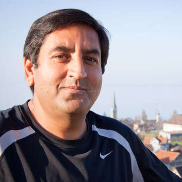 A man with dark hair is smiling slightly, wearing a black Nike sports shirt. He stands outdoors with a blurred cityscape and blue sky background.