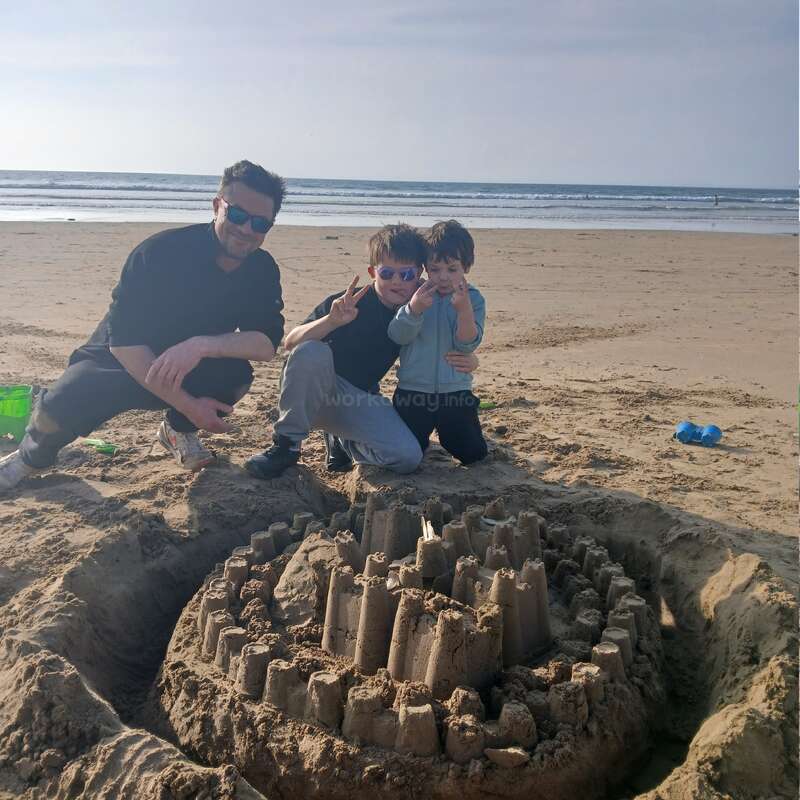 A man and two children kneel on a sandy beach beside a large, elaborate sandcastle. They smile and pose happily, enjoying a sunny, relaxing day together.