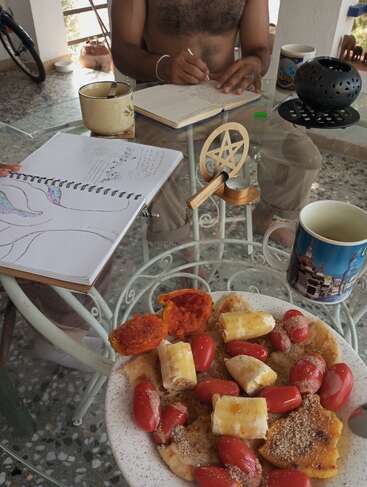 Auf einem Glastisch stehen Kaffeetassen, ein Teller mit Pfannkuchen mit Tomaten und Bananen, Skizzenbücher, ein Pentagramm-Kerzenhalter und zwei Menschen, die zeichnen und schreiben.