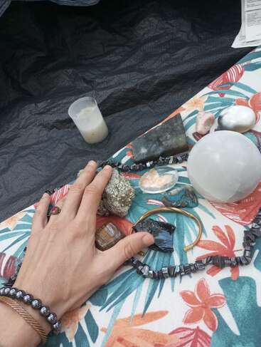 Eine Hand ruht auf einem bunten Blumentuch, das mit Kristallen, Steinen, Schmuck, einer Kerze und einer Quittung bedeckt ist und ein spirituelles und künstlerisches Arrangement bildet.