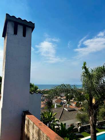 Im Vordergrund steht ein hoher weißer Schornstein, der den Blick auf Dächer, üppig grüne Palmen und ein entferntes, ruhiges Meer unter einem strahlend blauen Himmel freigibt.