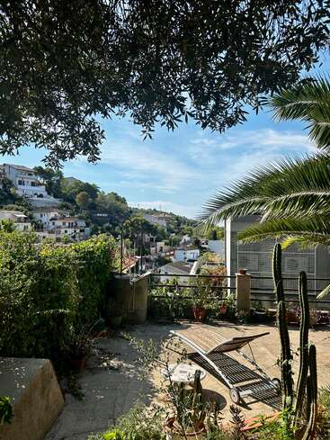 Eine sonnige Terrasse mit einem Liegestuhl, umgeben von verschiedenen Pflanzen und Kakteen. Mit Blick auf ein friedliches Dorf in den Bergen mit weißen Häusern, blauem Himmel und Bäumen.