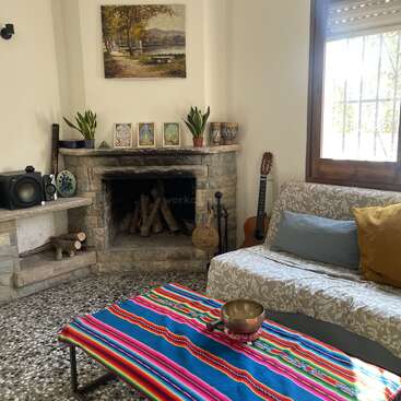 Ein gemütliches Wohnzimmer mit einem Steinkamin, Pflanzen, Kunstwerken, einer bunt gestreiften Tischdecke, einem Sofa, Kissen, einer Gitarre, einem Lautsprecher, einem Fenster und einer dekorativen Schale.