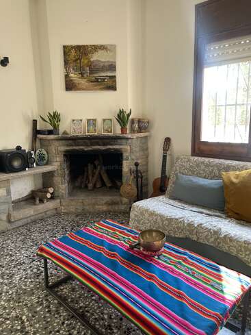 Ein gemütliches Wohnzimmer mit einem Steinkamin, Pflanzen, Kunstwerken, einer bunt gestreiften Tischdecke, einem Sofa, Kissen, einer Gitarre, einem Lautsprecher, einem Fenster und einer dekorativen Schale.