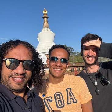Three smiling men take a selfie on a sunny day, standing in front of a white Buddhist stupa with a golden spire and clear blue sky.