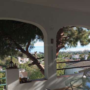 Eine sonnige, überdachte Terrasse mit weißen Bögen, einem Glastisch und Korbstühlen bietet einen Blick auf grüne Bäume, Dächer und das Meer in der Ferne und schafft so eine entspannte Atmosphäre.