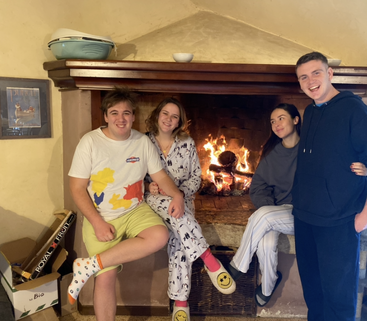 The image depicts a group of people gathered around a fireplace, with a table and chairs visible in the background, conveying a sense of warmth and togetherness.