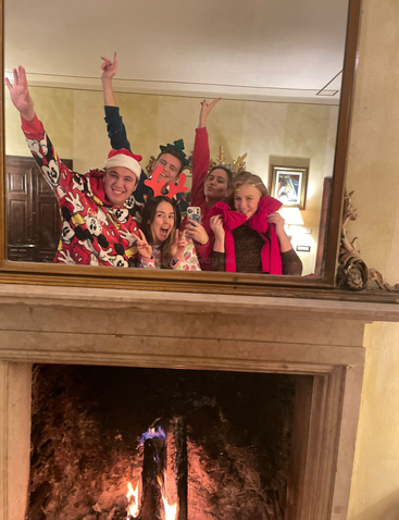 The image depicts a group of people gathered around a table, with a fireplace and a Christmas tree in the background, conveying a sense of warmth and festivity.